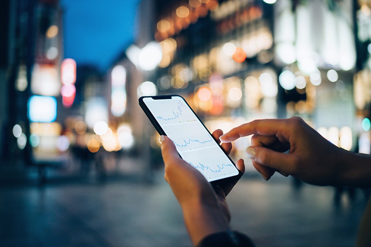 Empresas inovadoras de serviços financeiros estão usando as Communications APIs para transformar a experiência do cliente, encantando clientes às vezes hesitantes com autenticação em tempo real e interações por vídeo, voz e mensagens. Businesswoman reading financial trading data on smartphone in downtown city street against illuminated urban skyscrapers